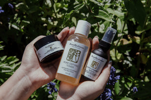 Hand holding three skincare products with a natural background of green leaves and purple flowers